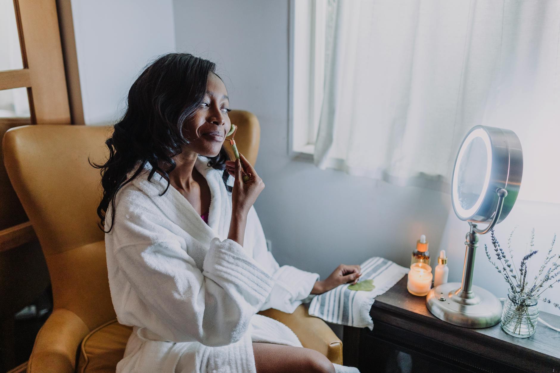 a woman in white bathrobe massaging her face with a jade roller
